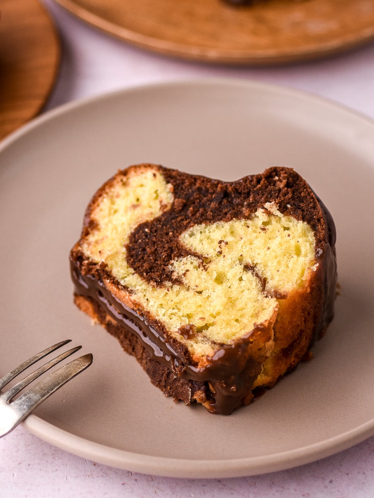 A slice of marble bundt cake shown on a plate. You can see the fluffy texture of the vanilla and chocolate cake as well as the shiny chocolate ganache coming down the side of the slice.