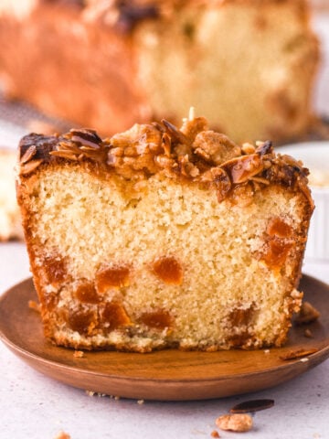 A slice of apricot and almond loaf cake on a small wooden plate. You can see the dried apricots dotted through the cake.