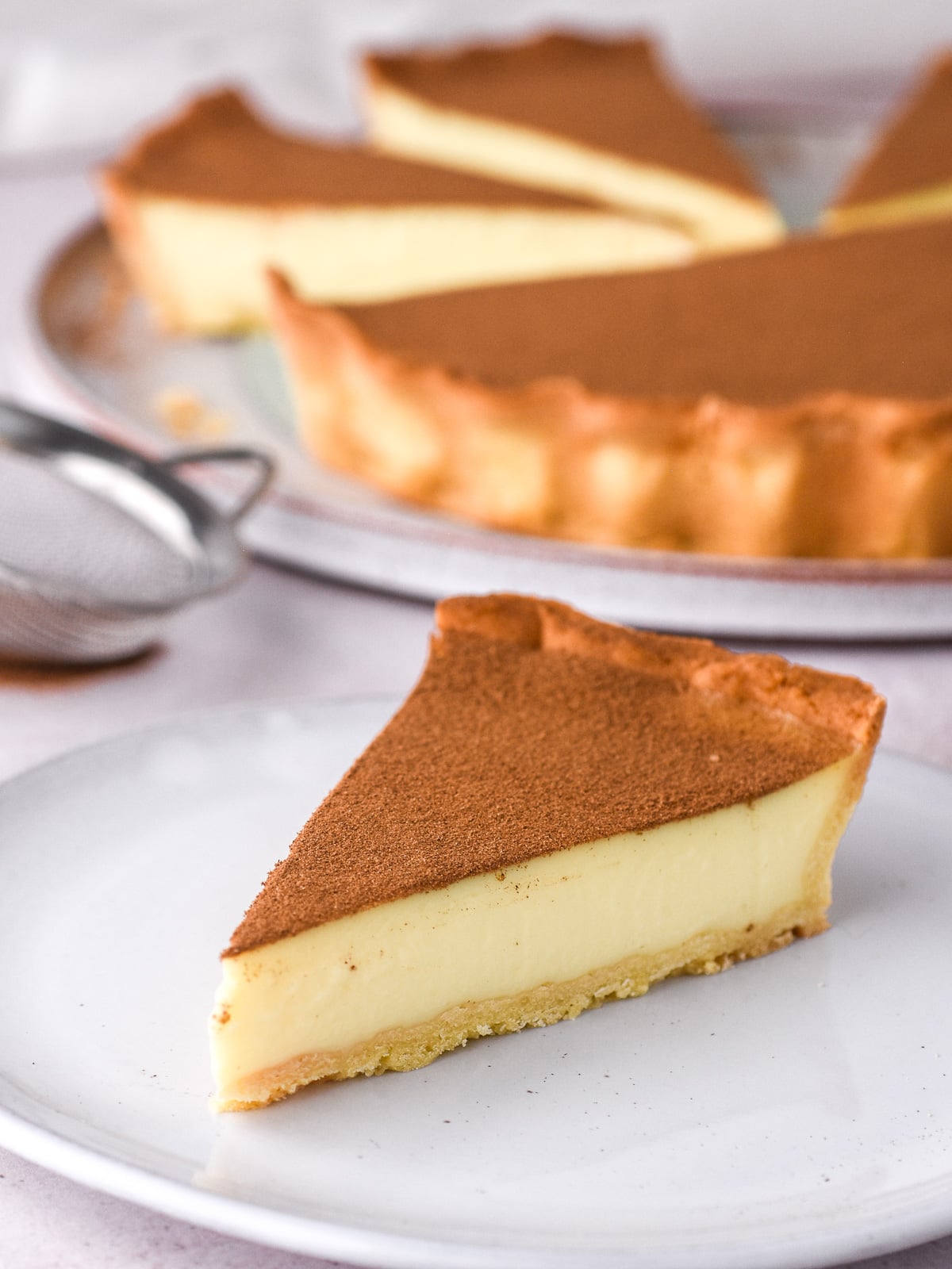 Image shows the perfect slice of milktart, the whole tart and a small sieve are in the background.