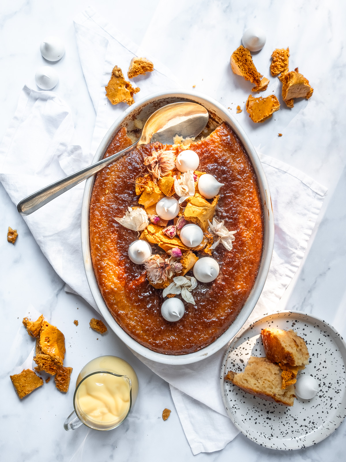 Malva pudding shown from above next to a jug of custard and crushed honeycomb pieces.