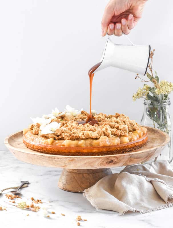 Apple Crumble Cheesecake shown on a cake stand with caramel sauce being poured onto it 