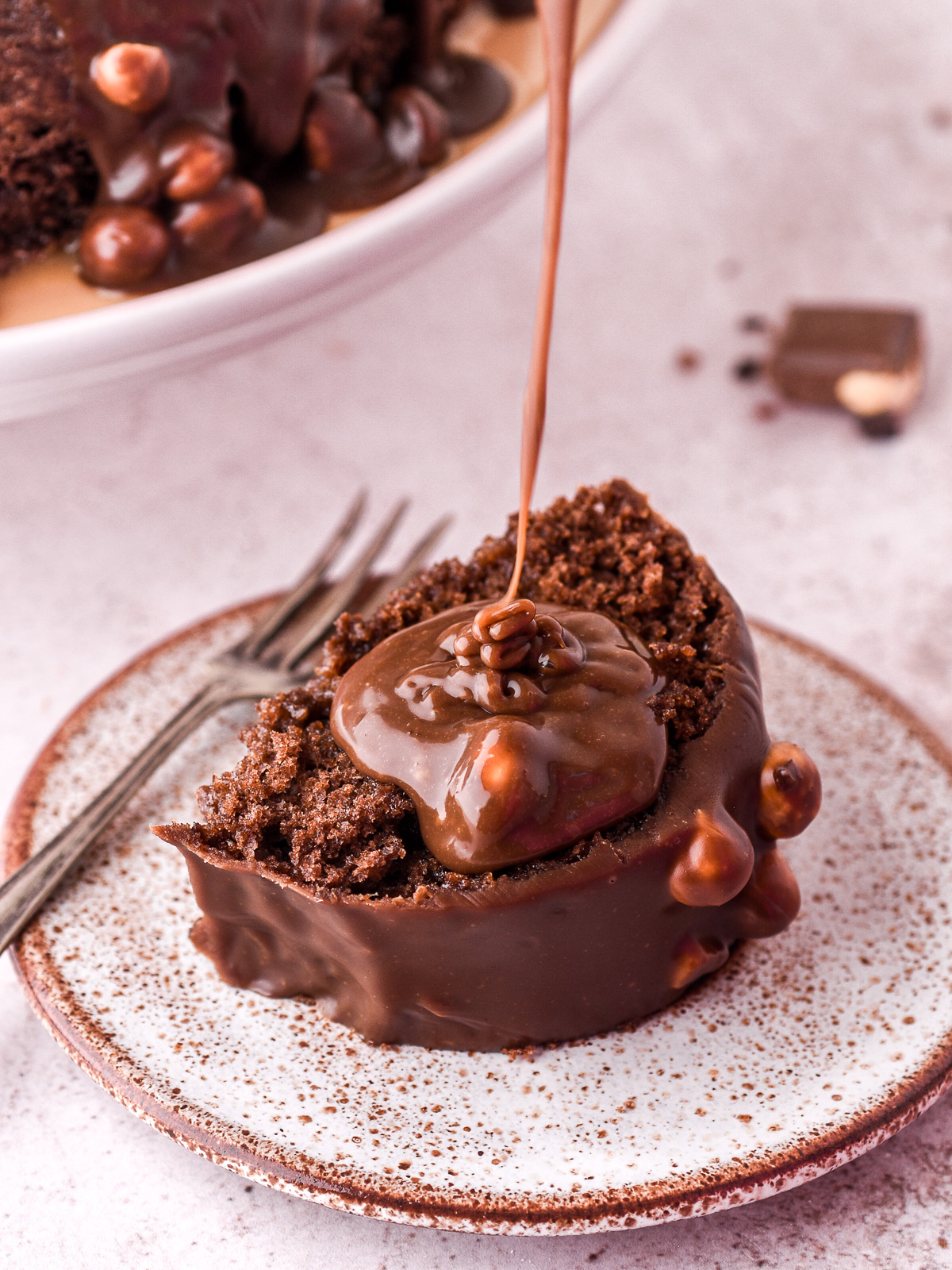 Image shows a slice of chocolate nut cake on a small plate, a drizzle of extra chocolate sauce is being poured over.