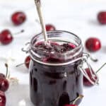 Image shows glass jar filled with cherry jam, there is a silver spoons ticking out of the jar.