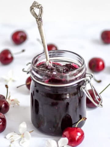 Image shows glass jar filled with cherry jam, there is a silver spoons ticking out of the jar.