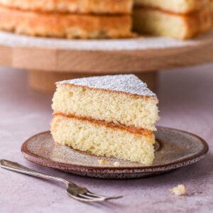 A slice of hot milk sponge cake shown on a small plate, the whole cake is in the background.