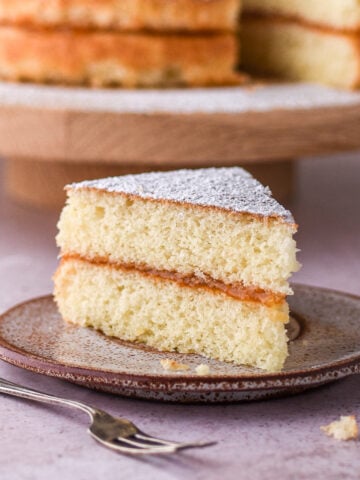 A slice of hot milk sponge cake shown on a small plate, the whole cake is in the background.