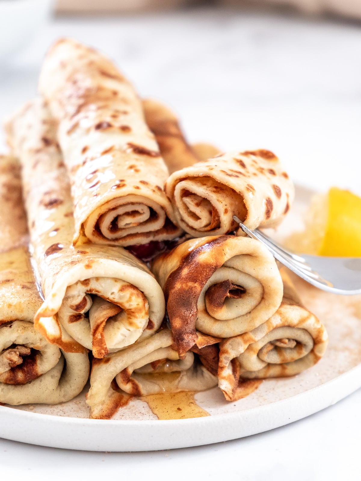 Image shows rolled up cinnamon sugar South African pancakes on a small white plate. A fork has cut through one of them.