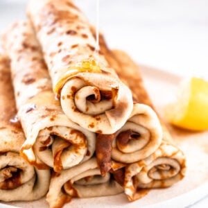 A stack of South African pancakes shown rolled up on top of each other. A drizzle of honey is coming down onto the pancakes.