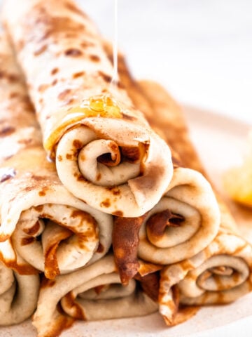 A stack of South African pancakes shown rolled up on top of each other. A drizzle of honey is coming down onto the pancakes.