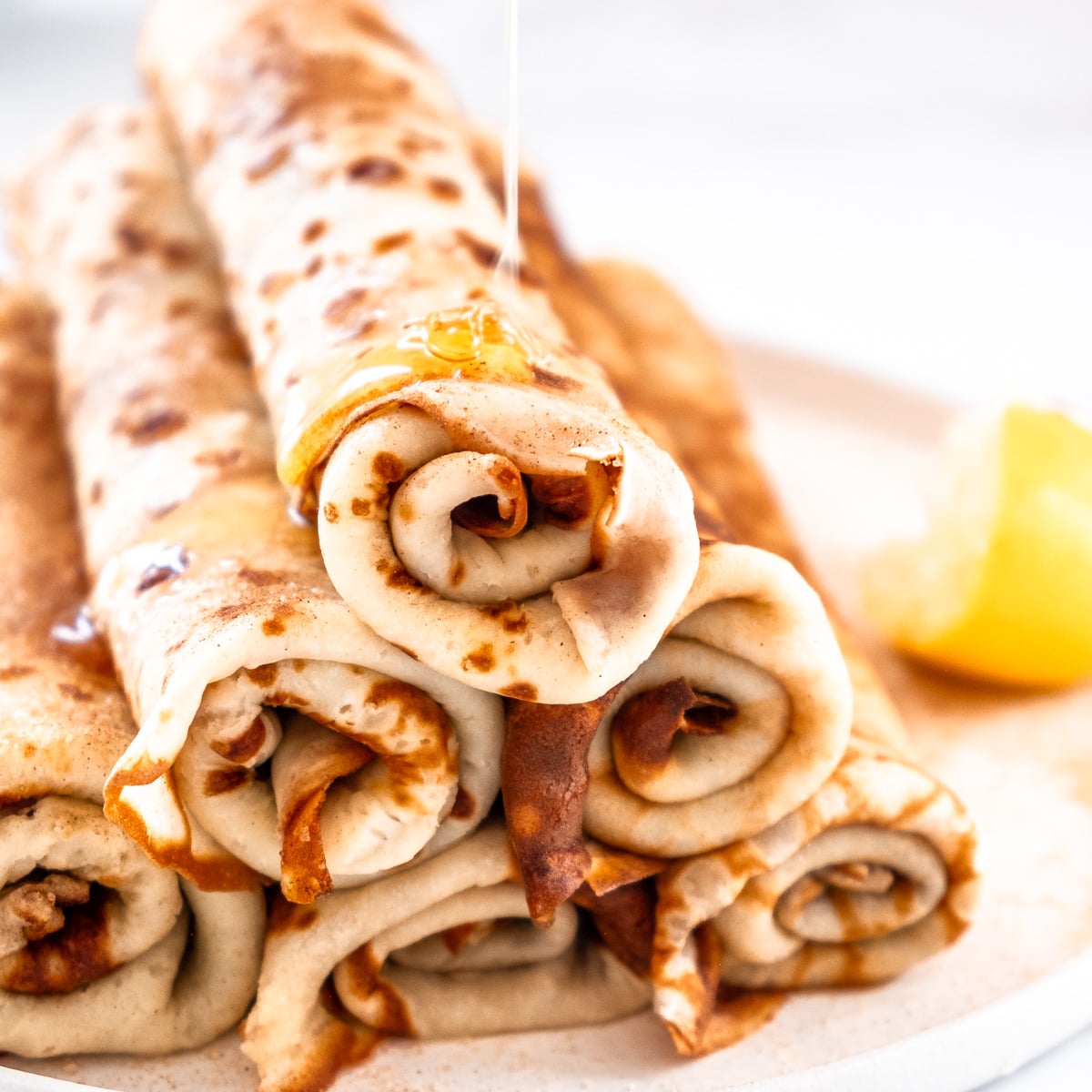 A stack of South African pancakes shown rolled up on top of each other. A drizzle of honey is coming down onto the pancakes.