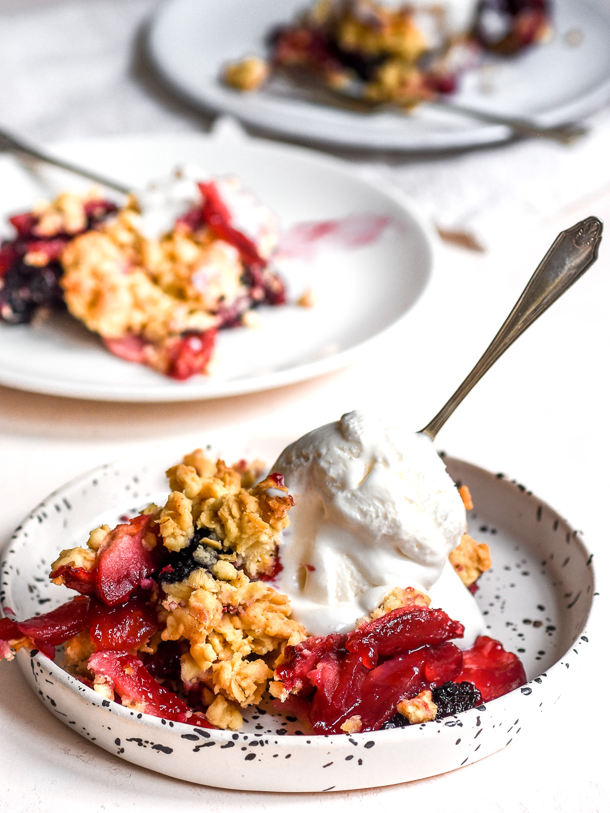 Apple and Blackberry Crumble shown on a plate with a scoop of ice cream. There are other plates in the background.