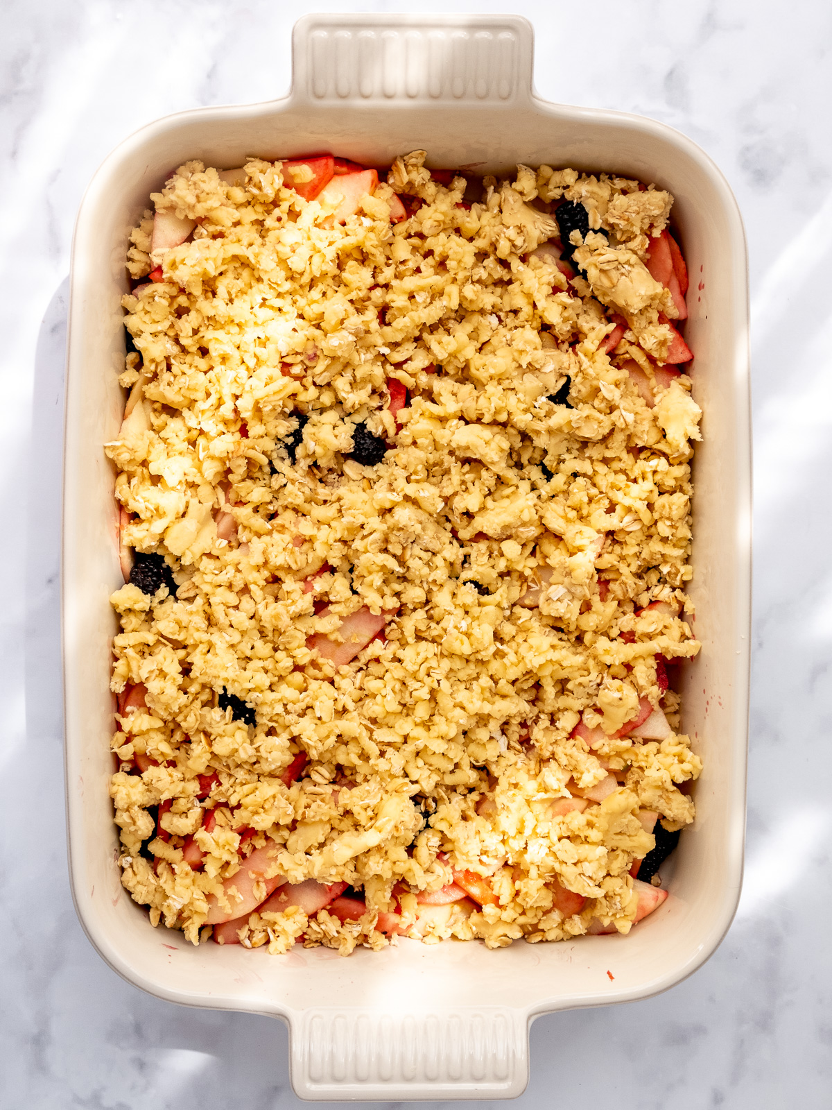 Image shows step 6 of how to make apple and blackberry crumble: grating the crumble dough over the fruit before baking.