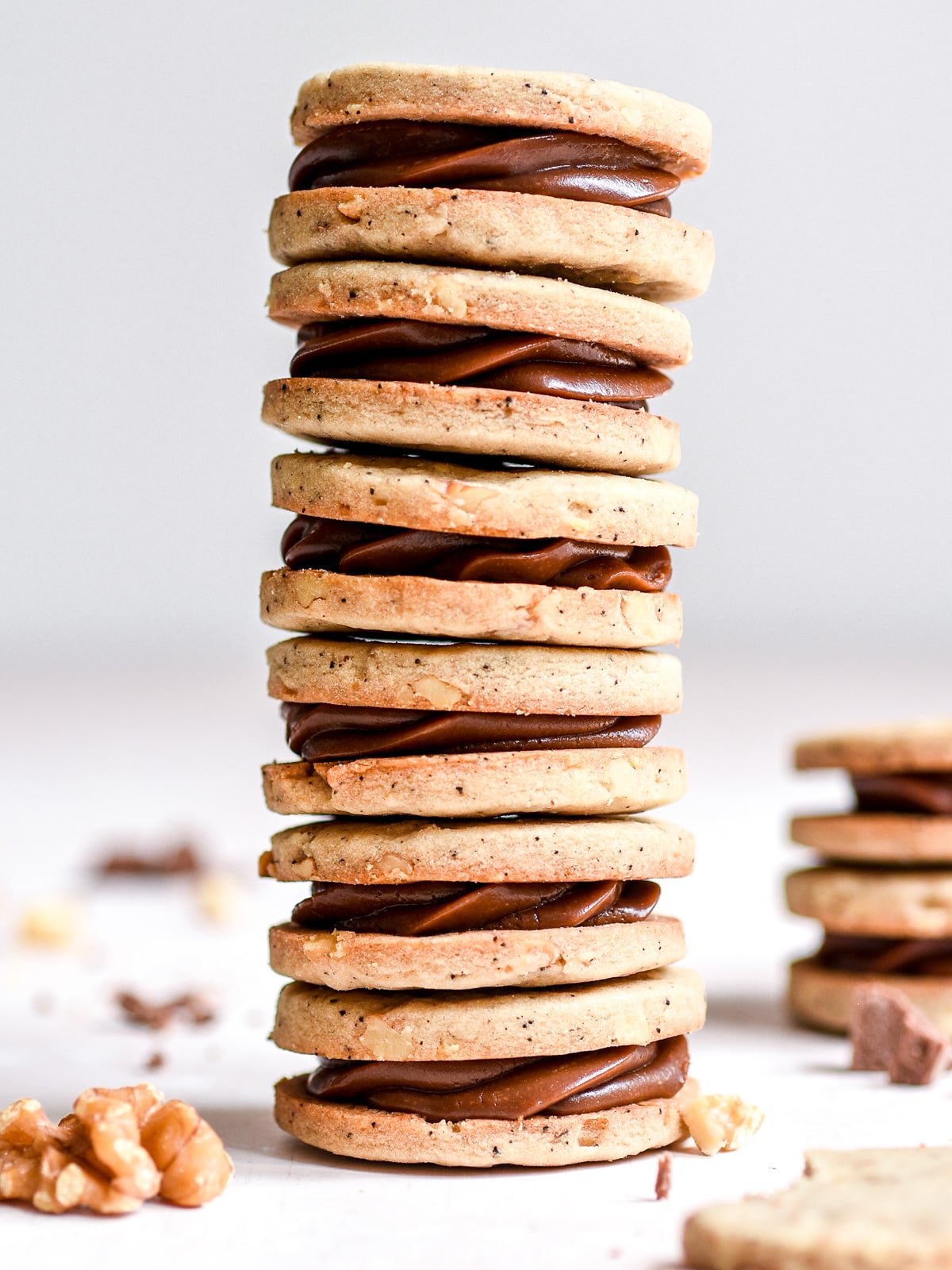Coffee and Chocolate Ganache Sandwich Cookies shown stacked on top of one another. There are walnuts and chunks of chocolate in the foreground.