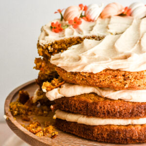 Image shows Pumpkin Carrot Cake with Brown Sugar Cream Cheese Frosting on a wooden cake stand with a slice missing.