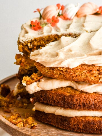 Image shows Pumpkin Carrot Cake with Brown Sugar Cream Cheese Frosting on a wooden cake stand with a slice missing.