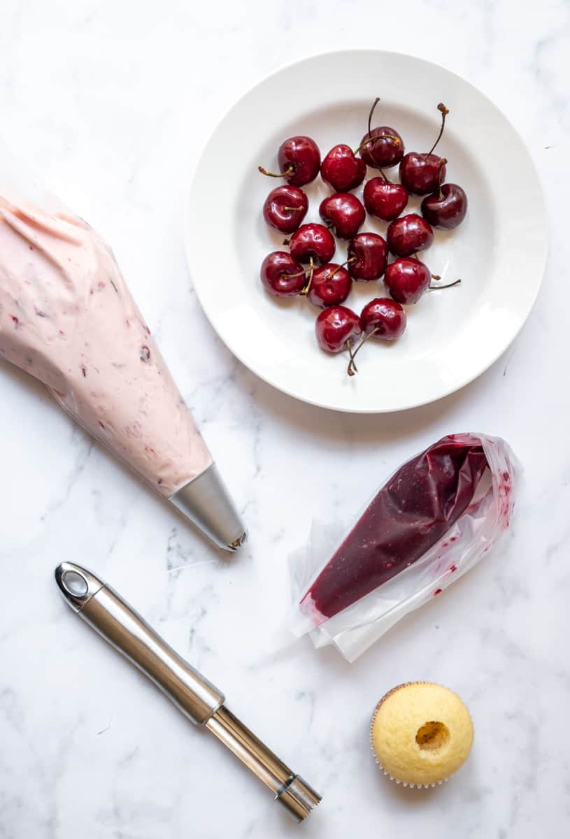 Photo showing a bowl of cherries, a piping bag filled with cherry frosting, a piping bag filled with the cherry filling and a cupcake with the cnter cored out. 