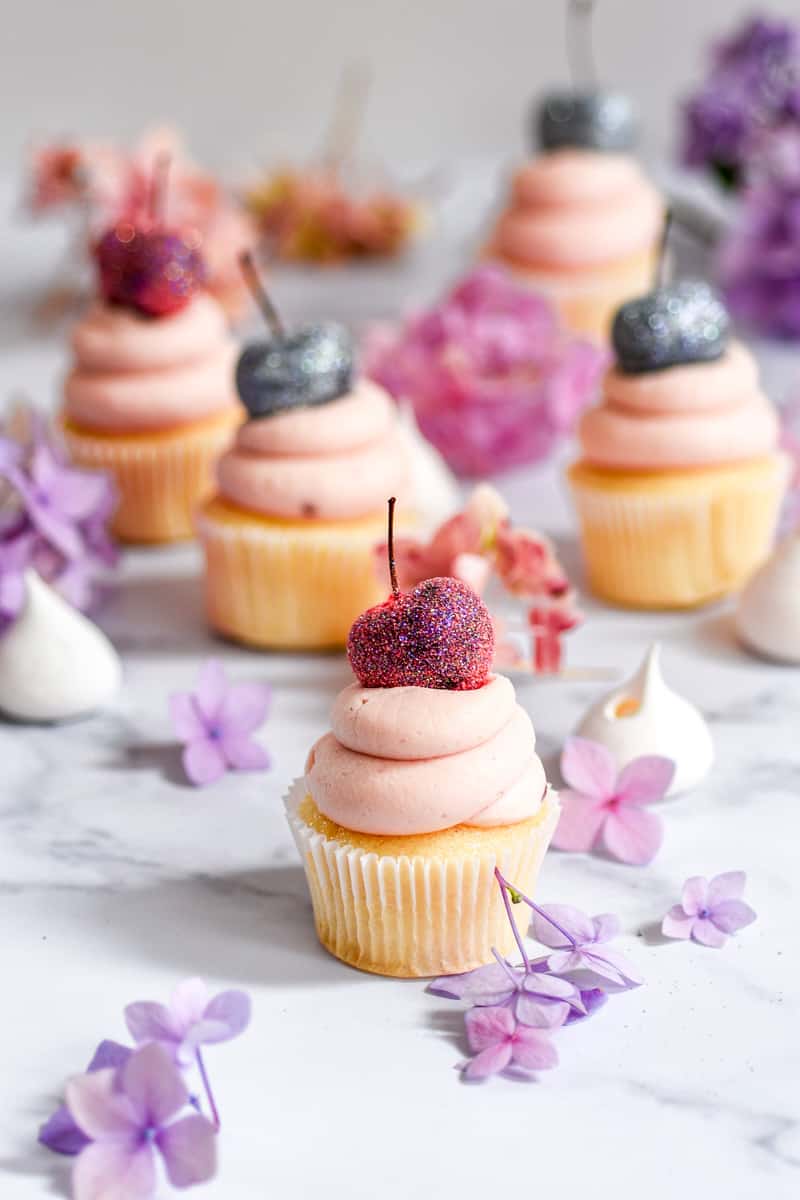 Cherry Frosting shown on mini vanilla cupcakes on a kitchen counter, with purple flowers.