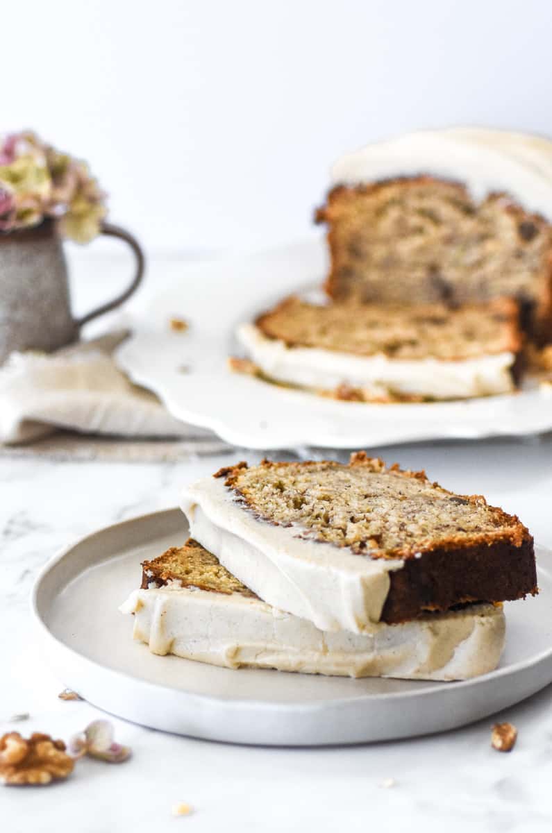 A plate with two slices on banana bread on it, with the loaf of the banana bread in the background. 