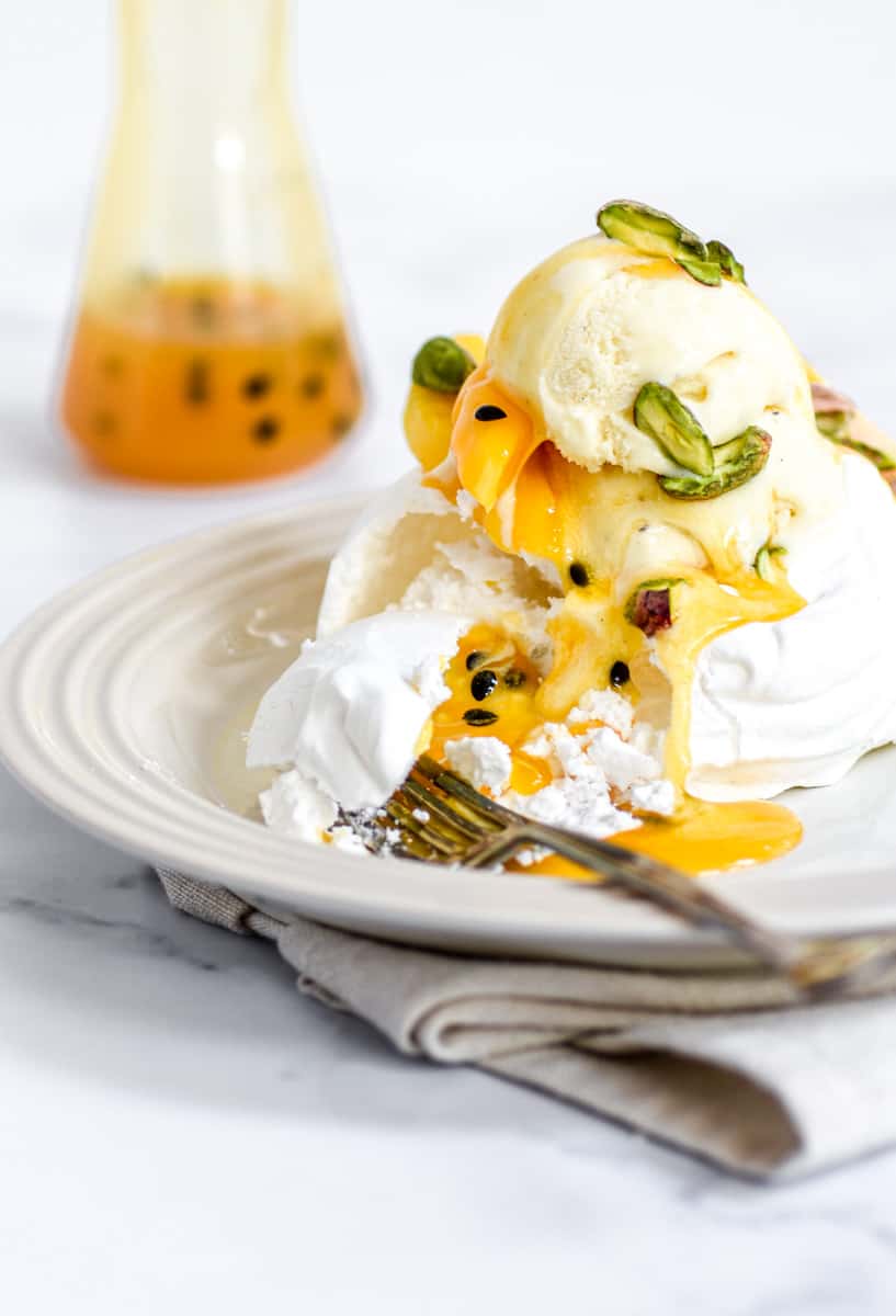 Mango Pavlova on a plate with a fork and a jar of passionfruit puree in the background.