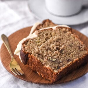 Image shows two slices of banana bread with cream cheese frosting on a small wooden plate.