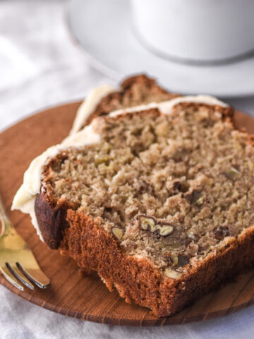 Image shows two slices of banana bread with cream cheese frosting on a small wooden plate.