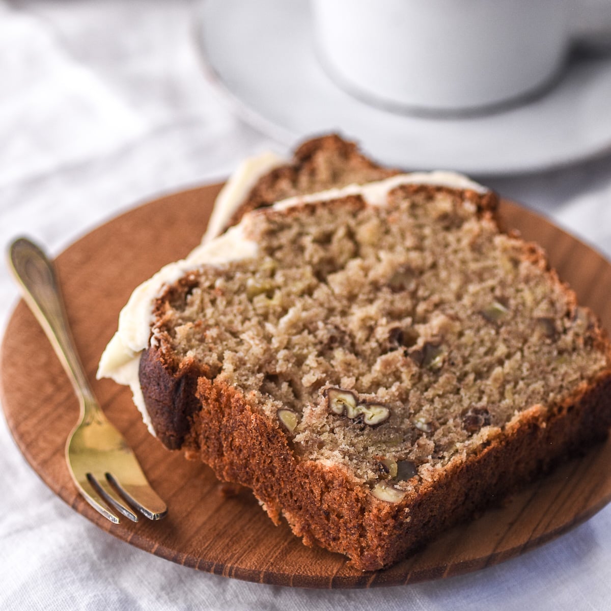 Image shows two slices of banana bread with cream cheese frosting on a small wooden plate.