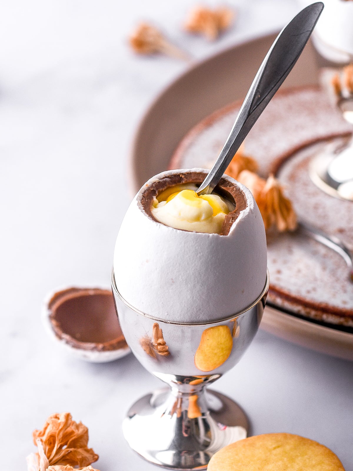 Image shows a candy coated Easter egg in a silver egg cup filled with cheesecake. There is a small spoon in the middle of the egg.