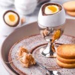 Image shows a cheesecake Easter egg in a silver egg cup next to biscuits and a small spoon.