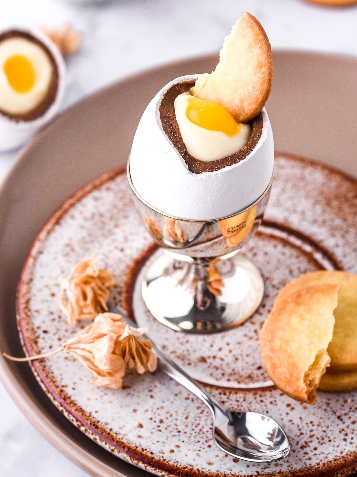 Image shows an Easter egg cheesecake in a silver egg cup with a biscuit through the middle of the mango 'yolk'.