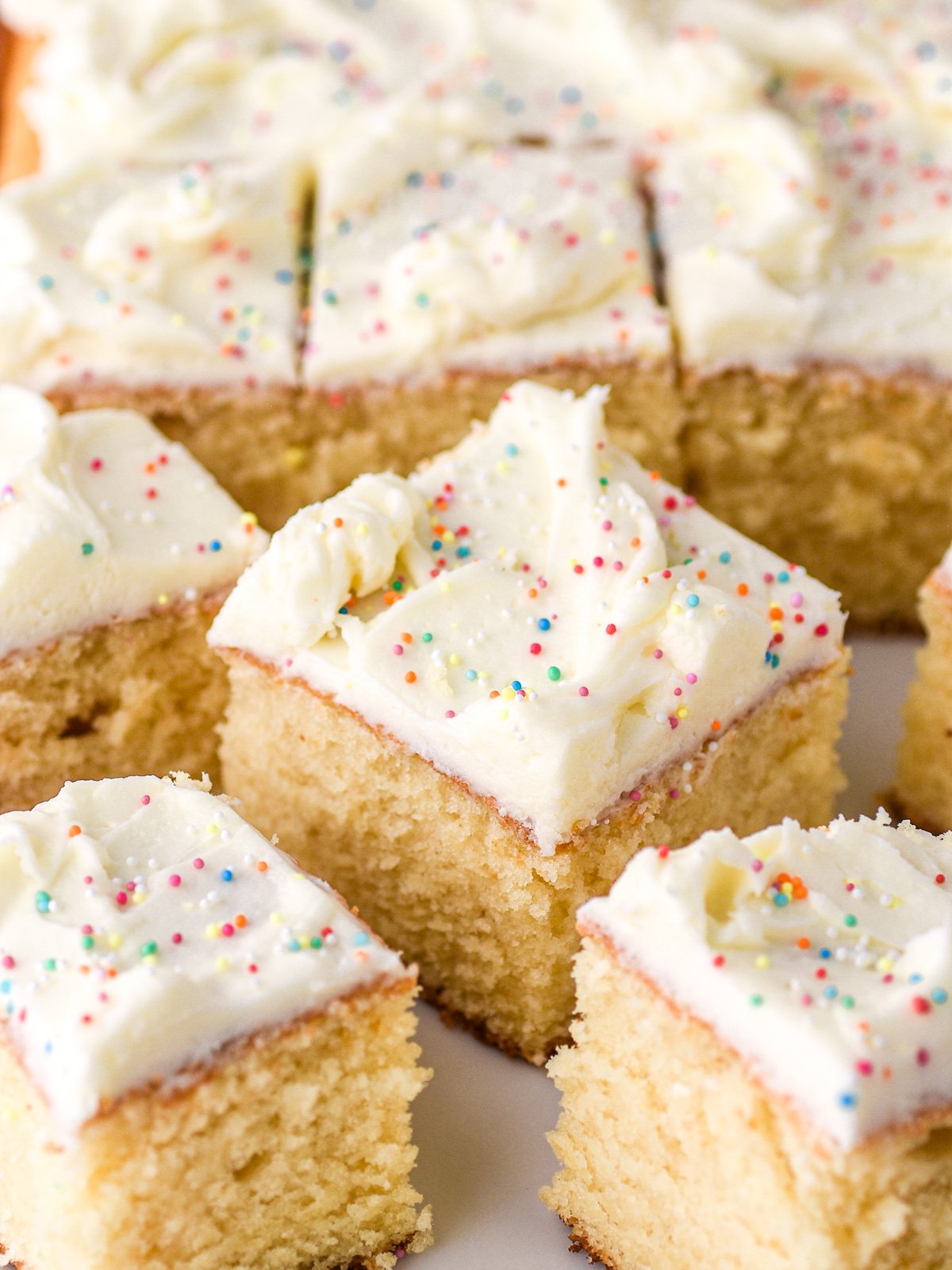 Image shows square slices of vanilla tray bake cake with vanilla buttercream frosting, the whole sheet cake is in the background.