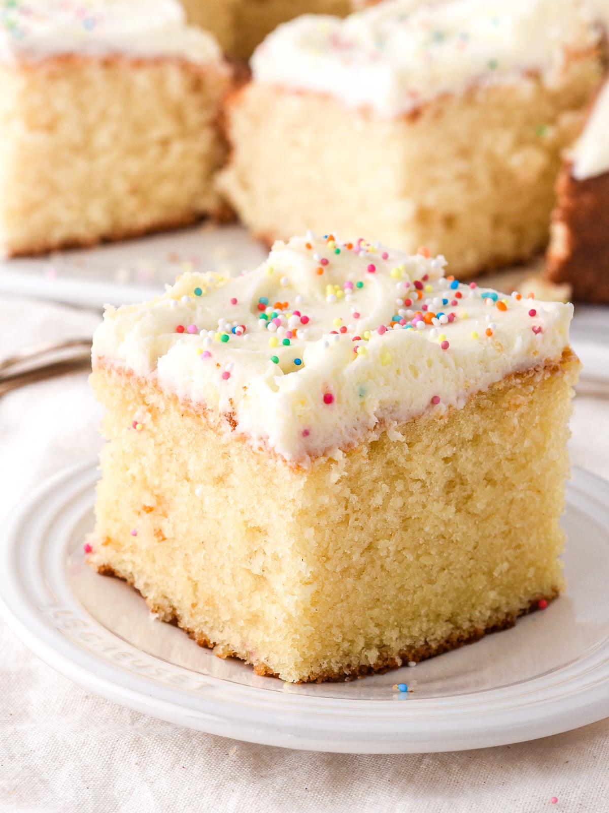 Square slice of vanilla tray bake cake with vanilla buttercream frosting and sprinkles shown on a small white plate.