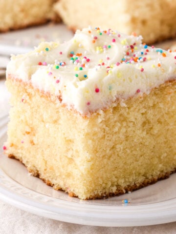 Square slice of vanilla sheet cake with vanilla frosting and sprinkles shown on a small plate.