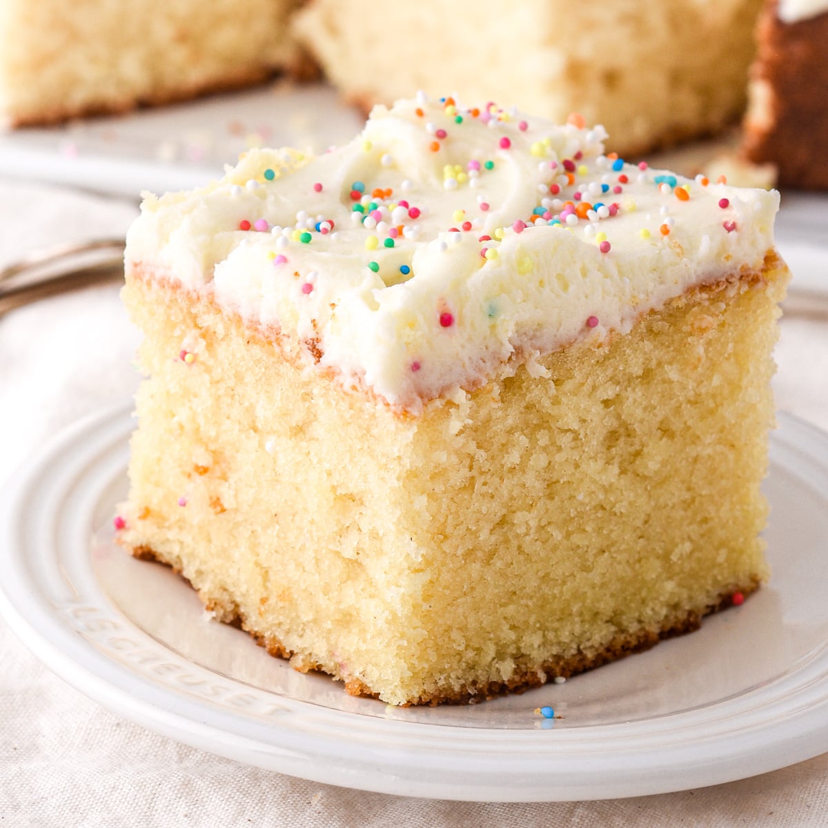 Square slice of vanilla sheet cake with vanilla frosting and sprinkles shown on a small plate.