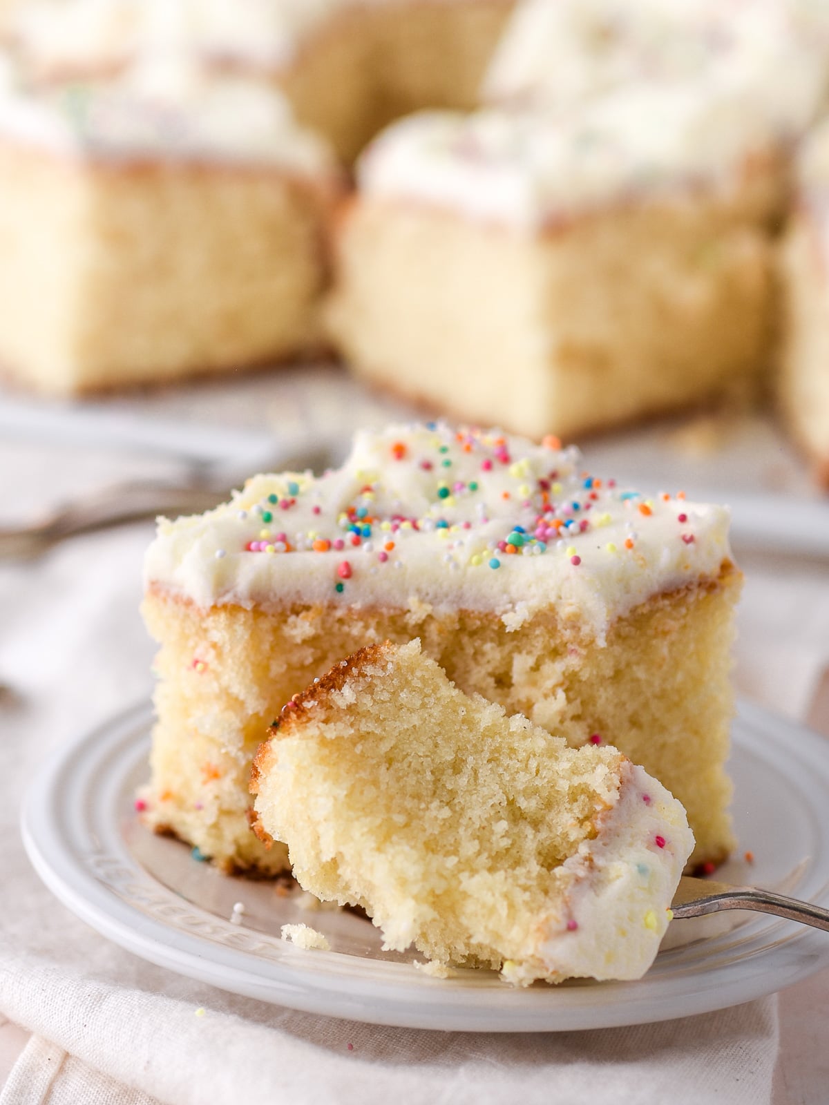 A slice of vanilla tray bake with vanilla buttercream shown on a small white plate, a fork has taken a bite away.