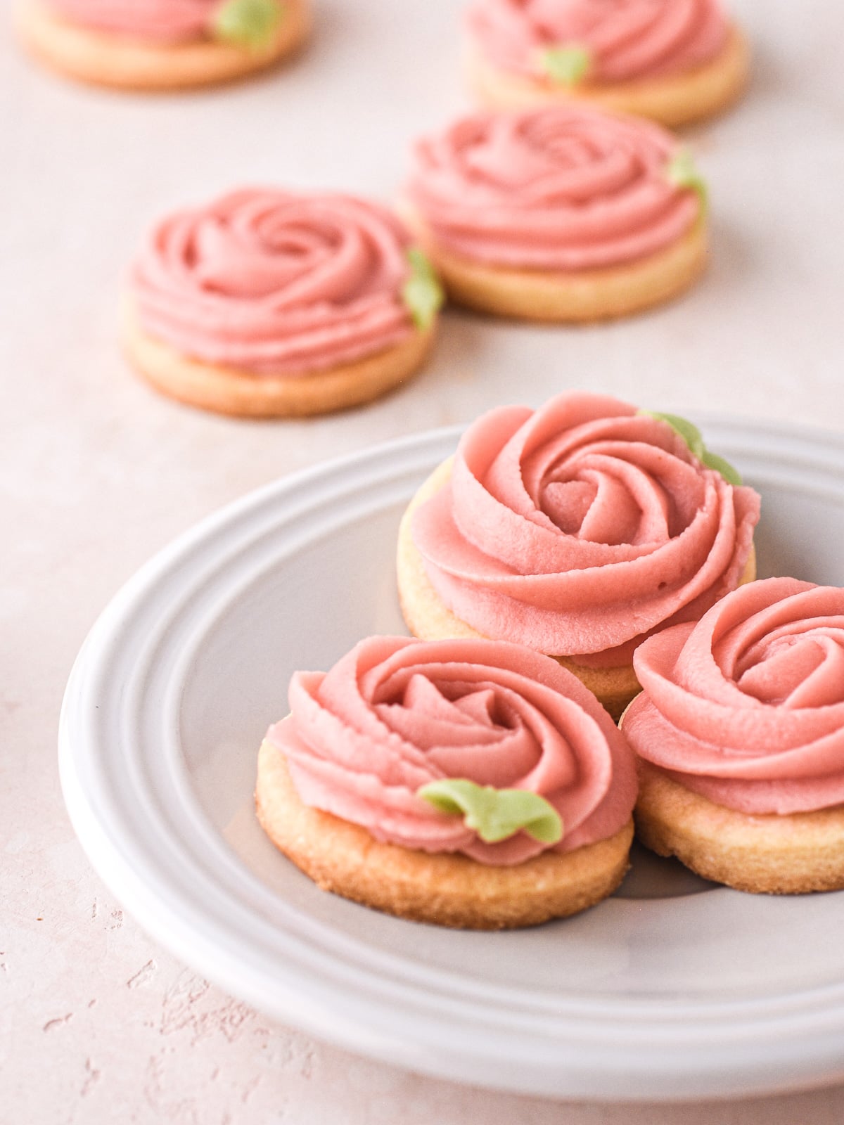 Image shows rose cookies on a white plate with extra cookies in the background.