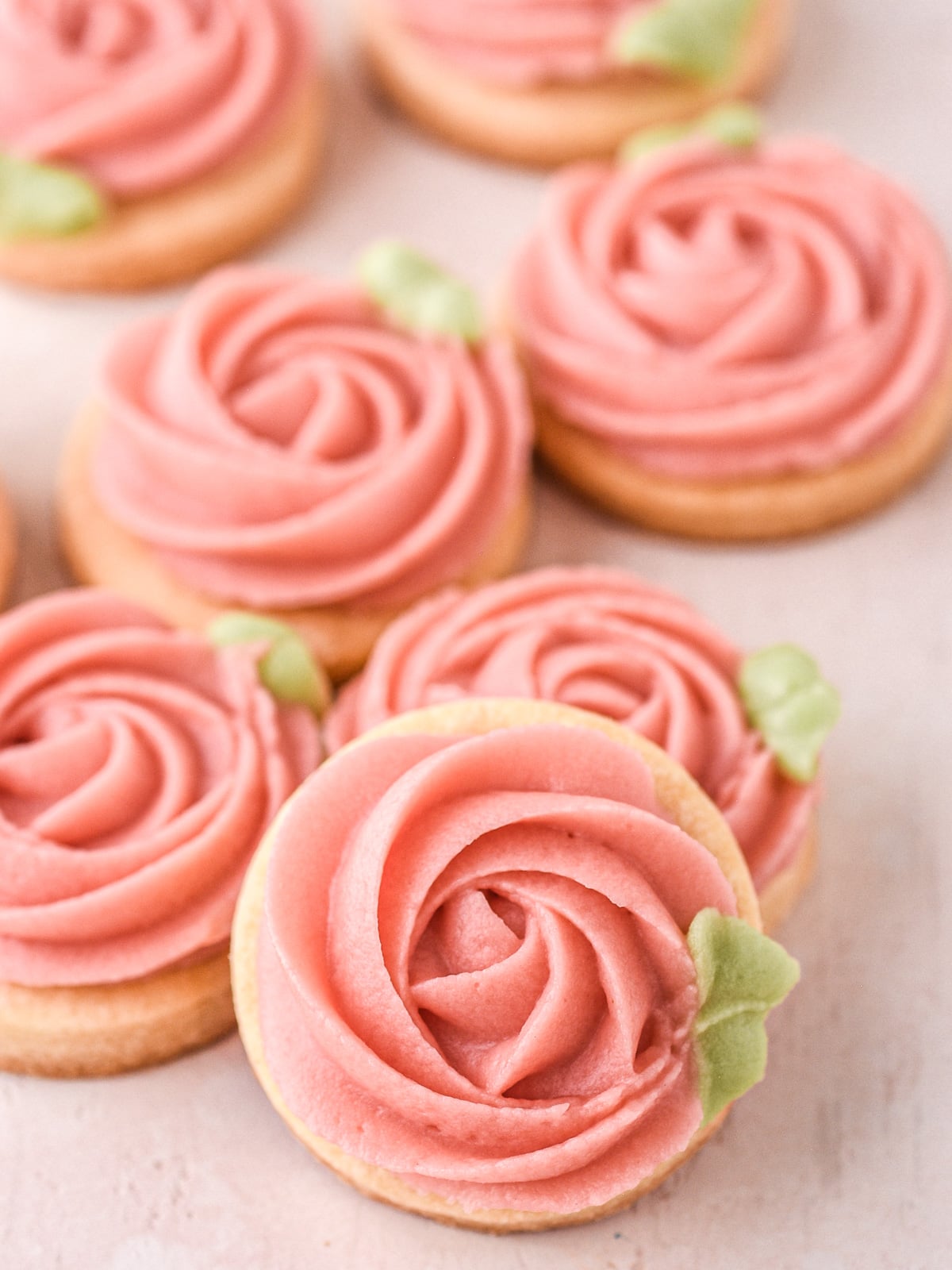 Image shows a few decorated buttercream cookies on a counter at different angles so that you can see the piping of the rose shape.