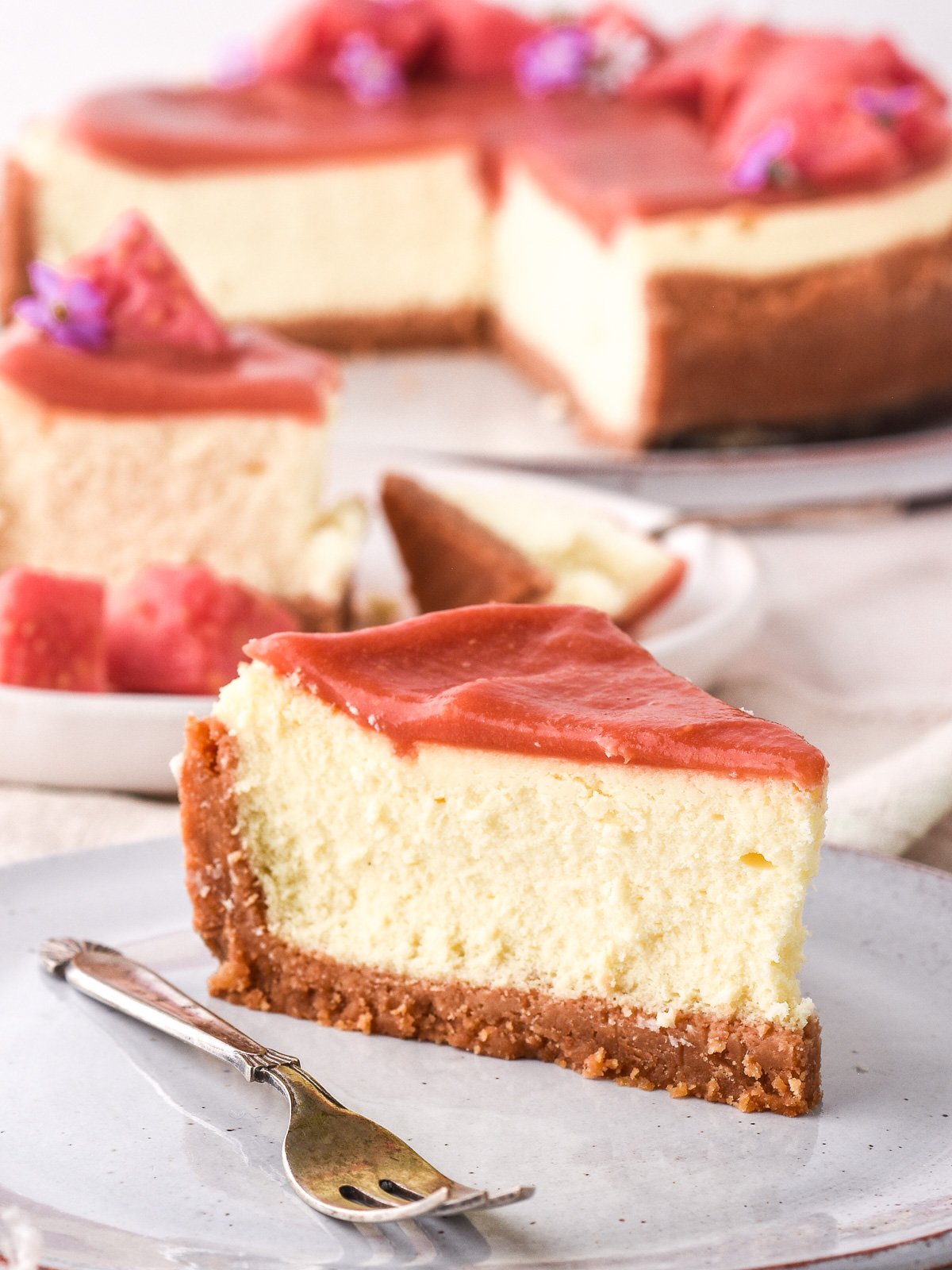 Image shows a slice of the cheesecake in the foreground with other slices and the rest of the cheesecake in the background.