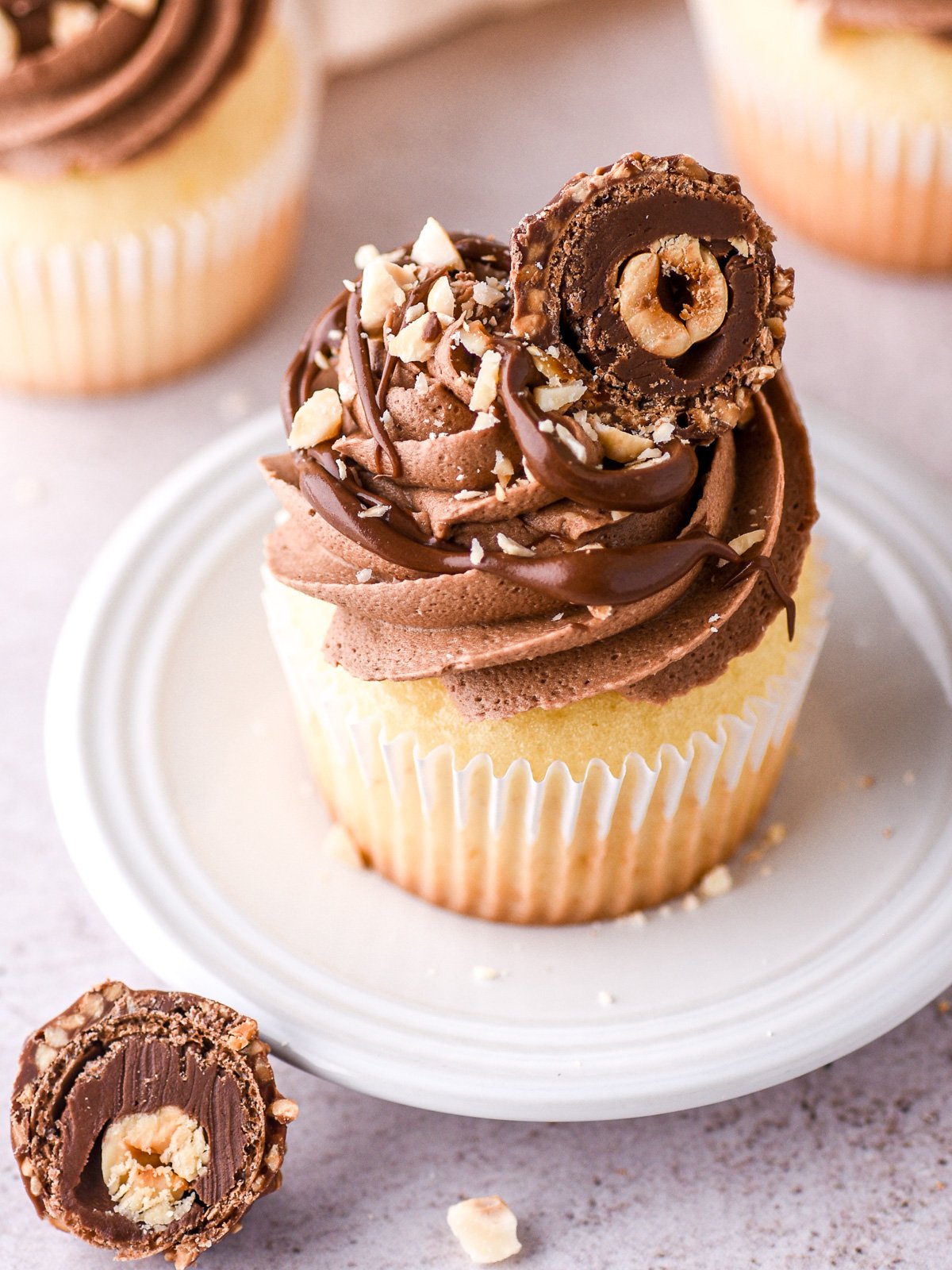 Image showing a vanilla cupcake swirled with Nutella buttercream frosting. It is decorated with chopped hazelnuts and a halved Ferrero Rocher.