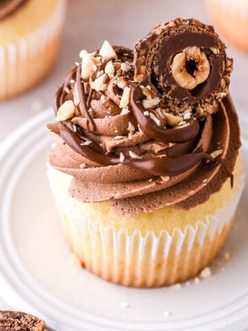 Vanilla Nutella Cupcake shown on a small white plate, it is decorated with chopped nuts and half a Ferrero Rocher Chocolate.