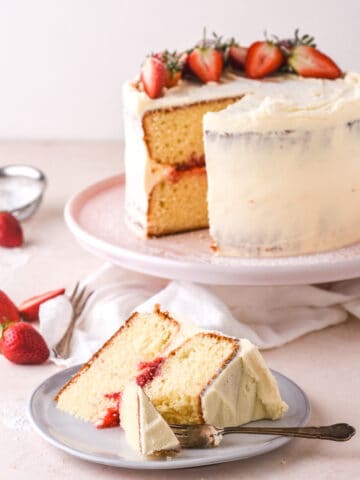 Vanilla with strawberry filling cake shown on a kitchen counter. There is a slice taken from the cake, this is shown on a plate in front of the cake.