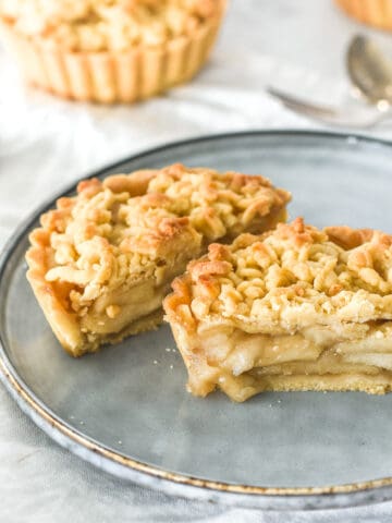 Mini apple crumble tarts shown on a kitchen counter. The one tart is cut in half and seen on a dusty blue plate.