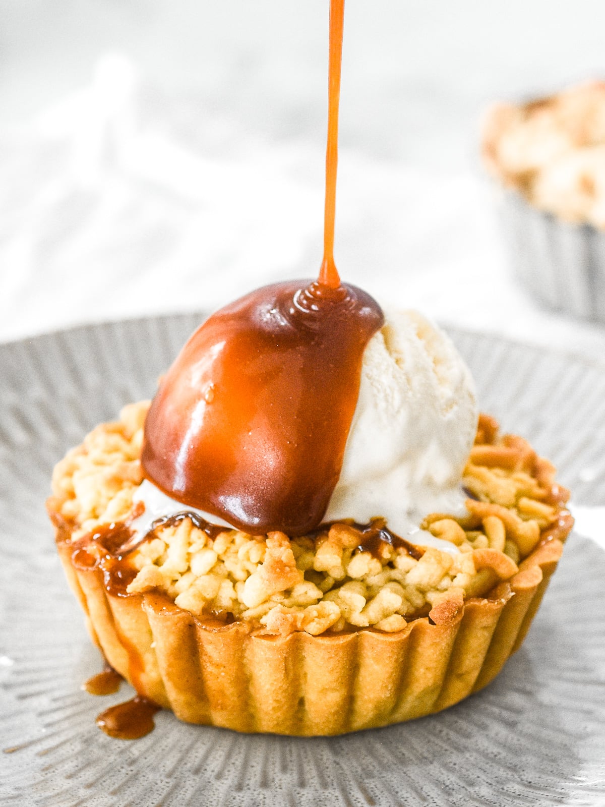 Apple crumble tartlet shown with a scoop of vanilla ice cream on top of the tart and a drizzle of caramel being poured over.