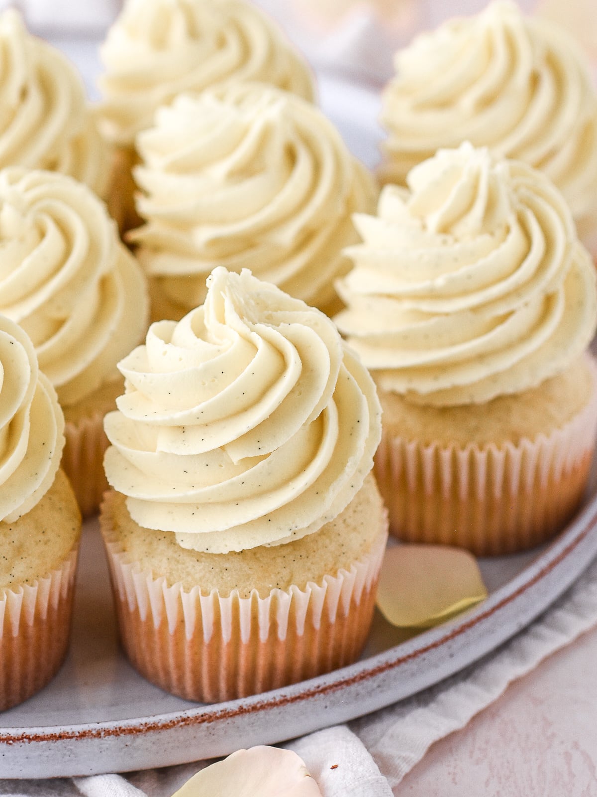 Image shows vanilla cupcakes piped with tall swirls of vanilla bean buttercream. You can see the black vanilla seeds in the frosting.