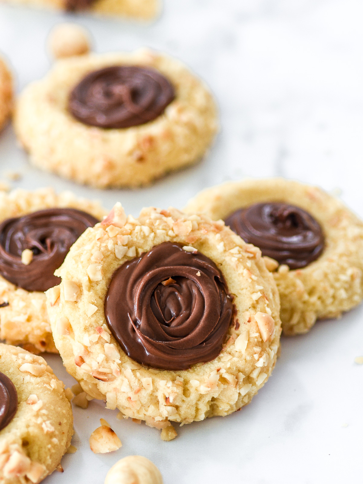 Nutella Thumbprint Cookies shown on a counter top next to crushed and whole hazelnuts.