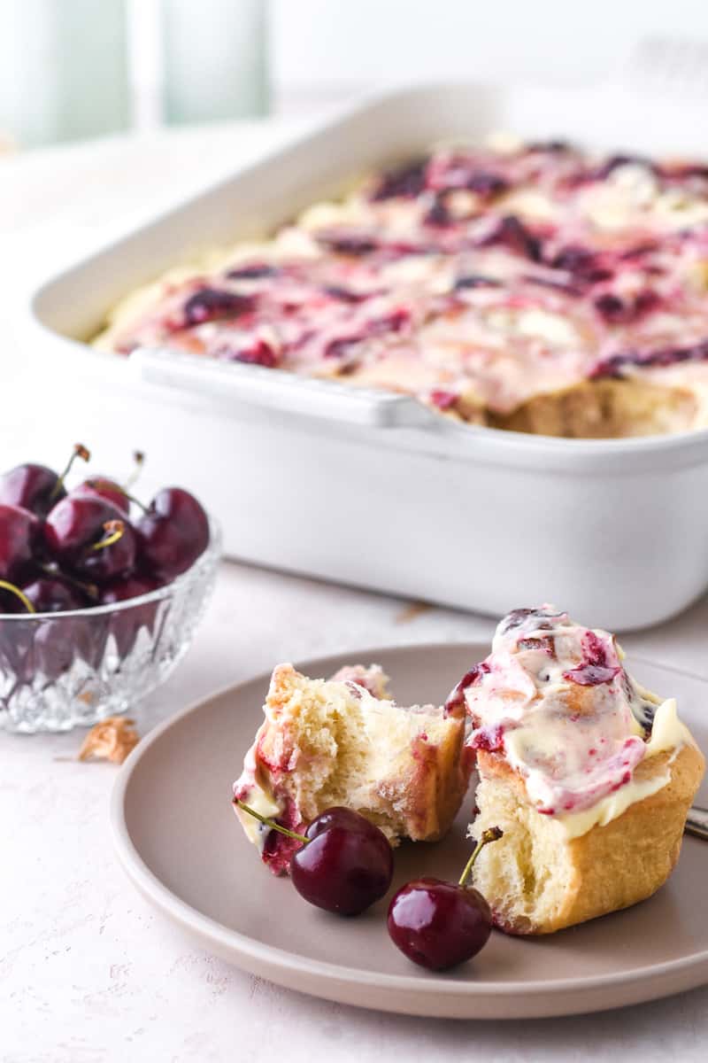 Cherry Cinnamon Roll shown on a plate next to a bowl of fresh cherries.