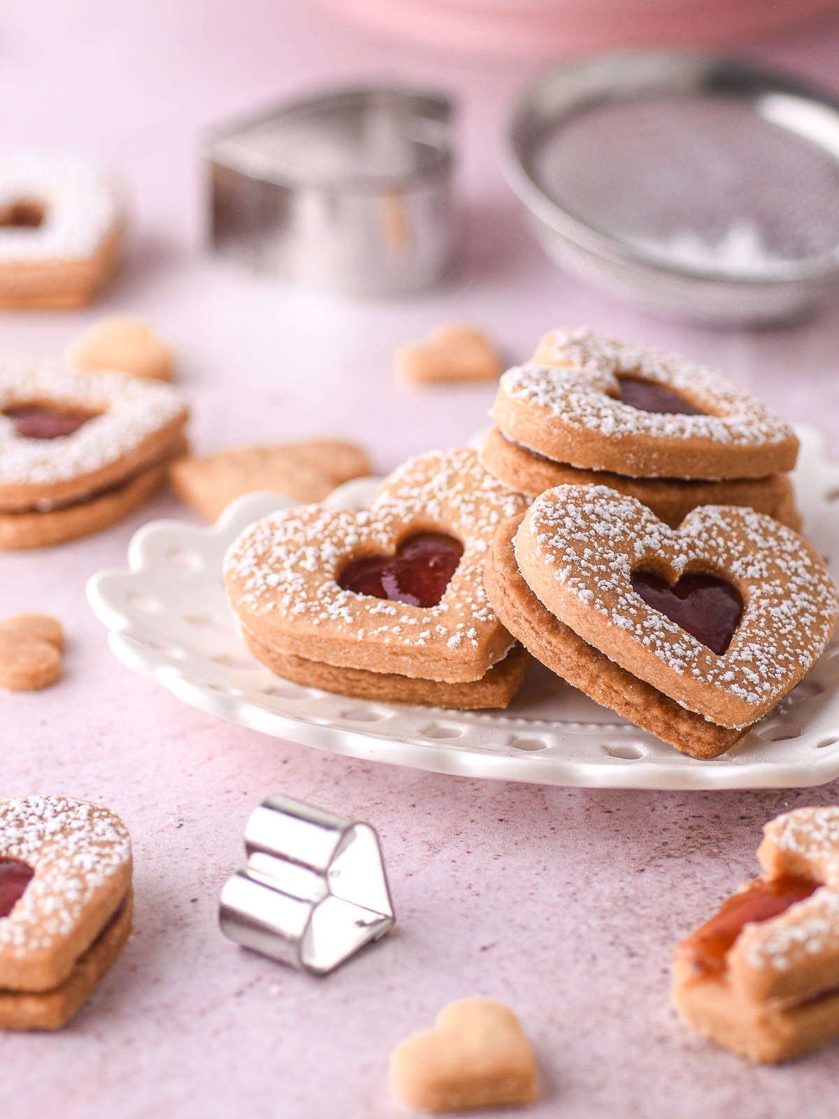 Image shows cookies on a white plate, there are cookies, cutters, and a sieve in the background.