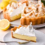Biscuit base lemon meringue pie shown on a kitchen counter, there is a slice missing from the pie and this is shown in the foreground of the image on a plate.