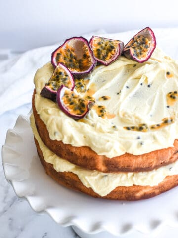 Passion Fruit cake shown on a white cake stand on a counter top. There are fresh passionfruit slices on top of the cake.