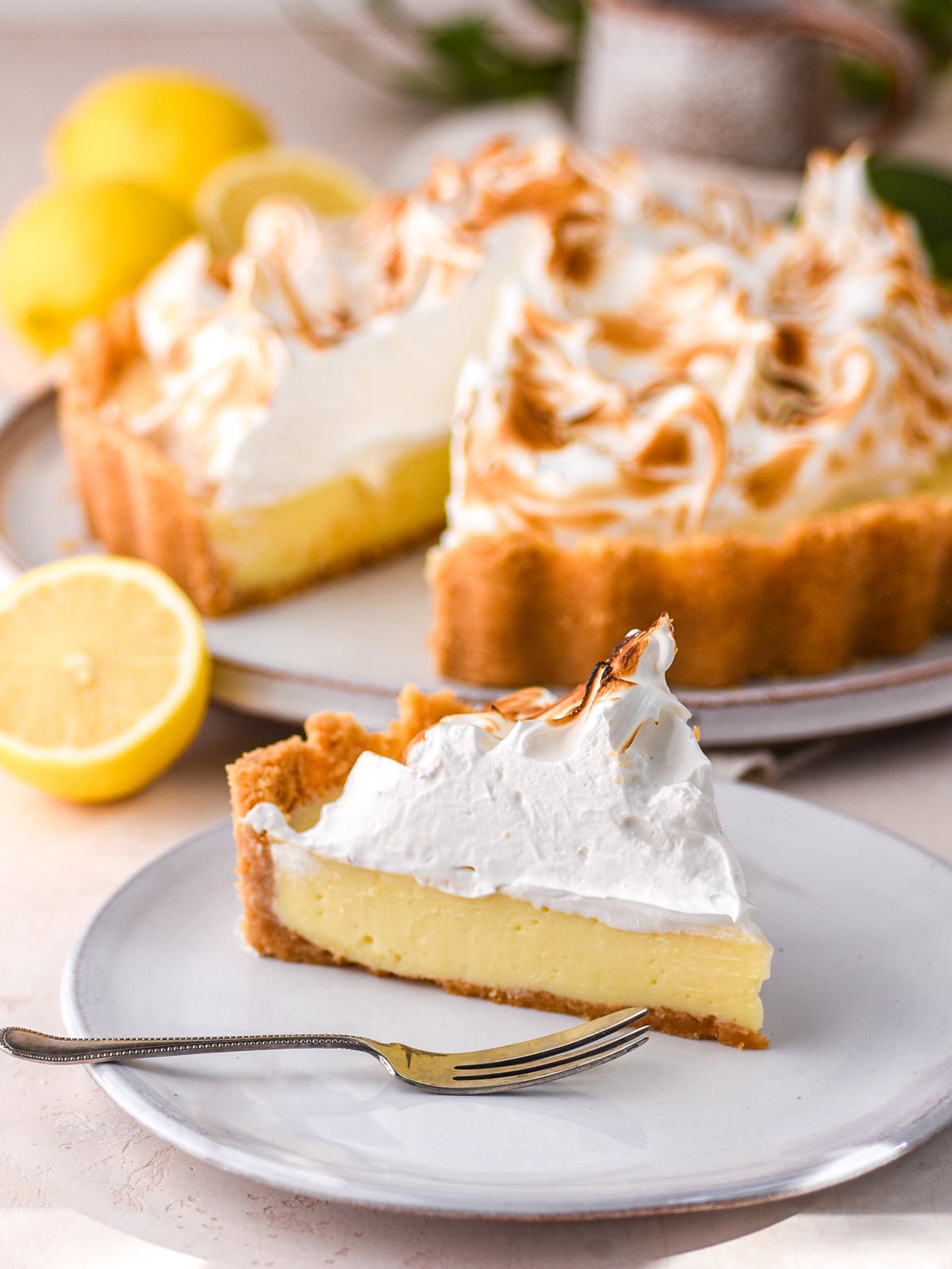 A slice of lemon meringue pie shown on a side plate, the whole pie and fresh lemons are seen in the background.