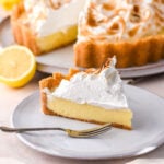 A slice of lemon meringue pie shown on a plate, the whole pie is in the background.
